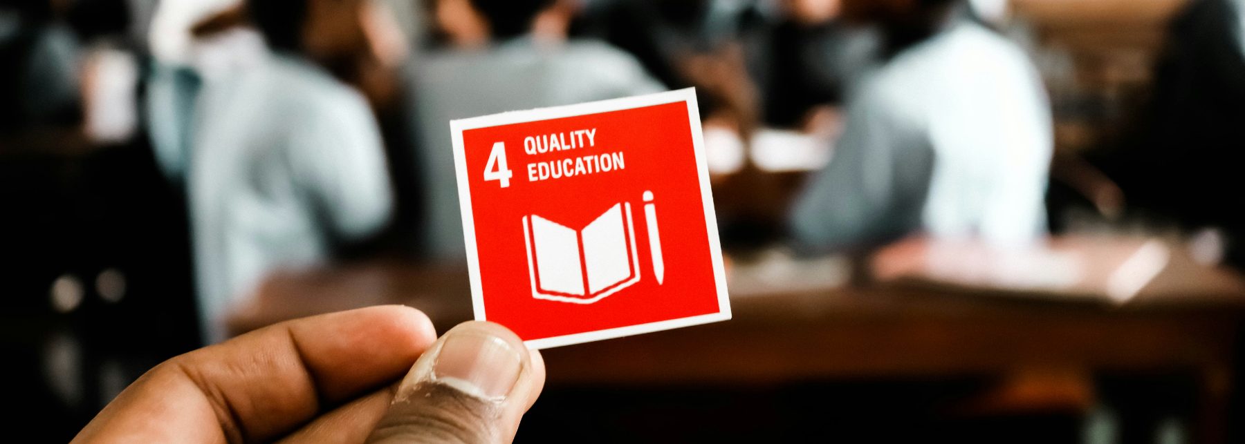 A hand holding the Sustainable Development Goal 4 card, Quality Education, in front of a classroom